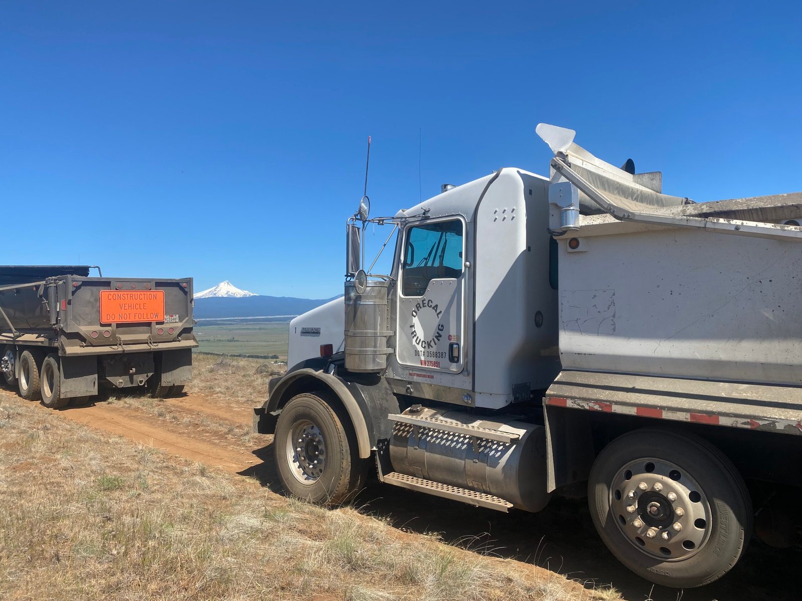 Orecal Trucking dump trucks on a rural dirt jobsite road with Mount McLoughlin in the background.